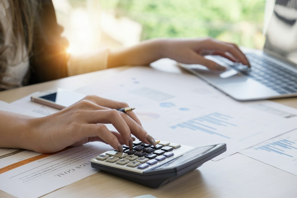 Women&rsquo;s hands at a desk typing into a calculator and a laptop.